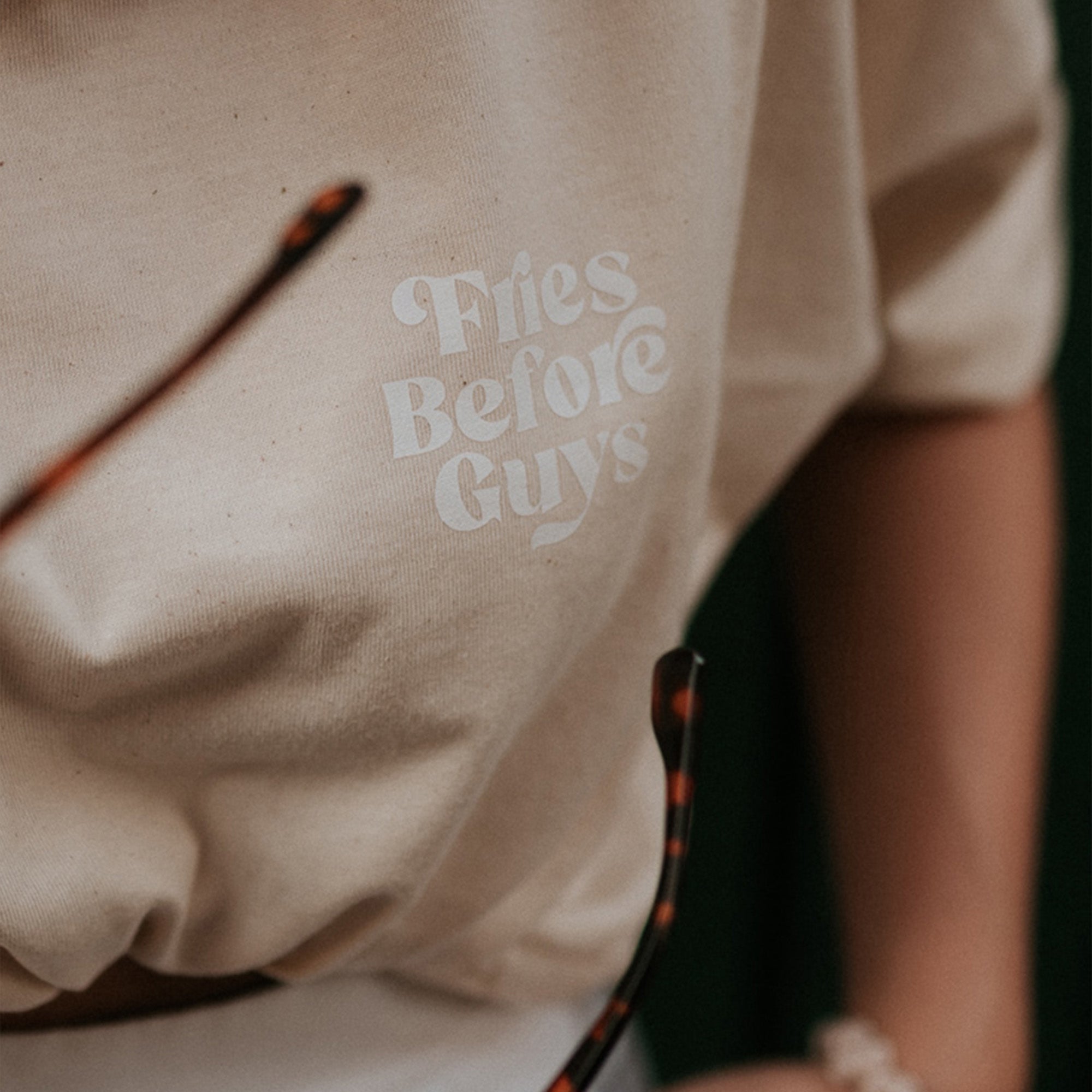 Detailaufnahme von weißem "Fries Before Guys"-Drucks auf der Vorderseite eines vanillafarbenen T-Shirts