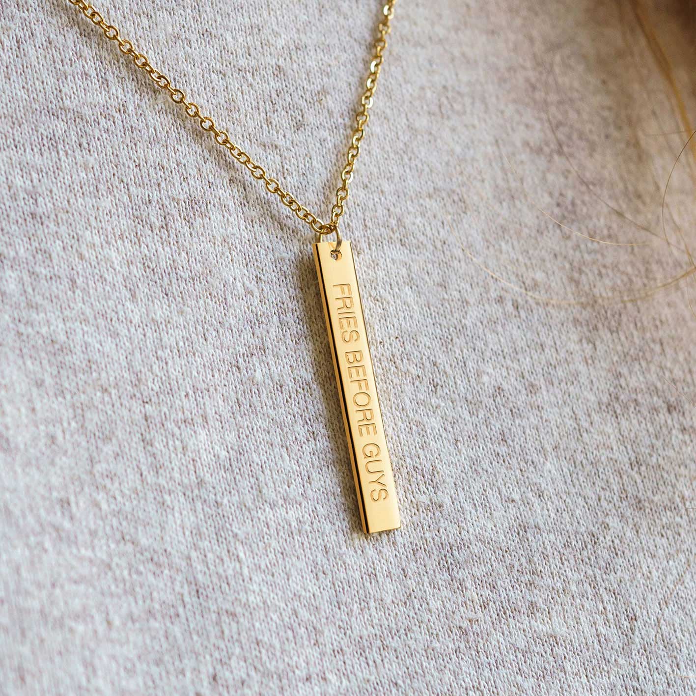 Detailansicht einer goldenen Kette mit länglichem Anhänger und "Fries Before Guys"-Gravur.