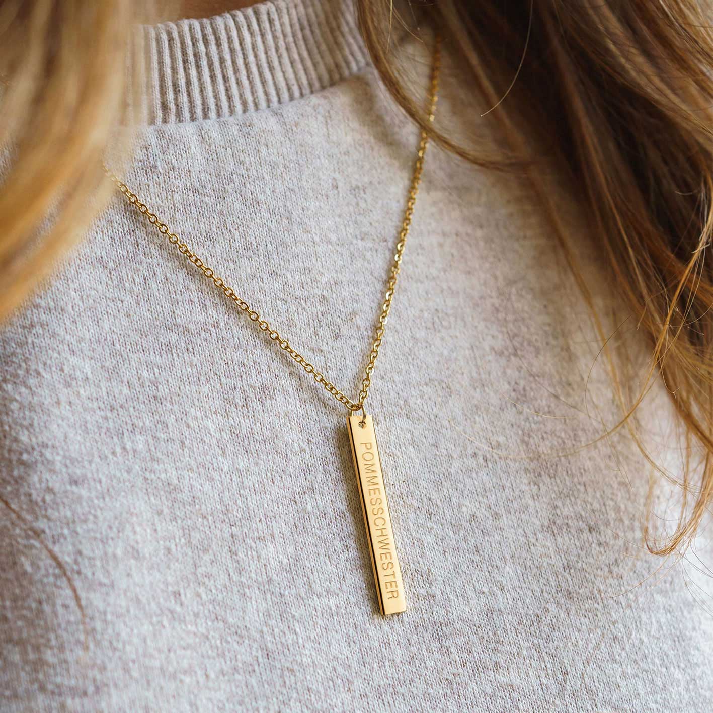 Frau mit langen Haaren trägt goldene Kette mit länglichem Anhänger mit "Pommesschwester"-Gravur auf beigen Pulli.