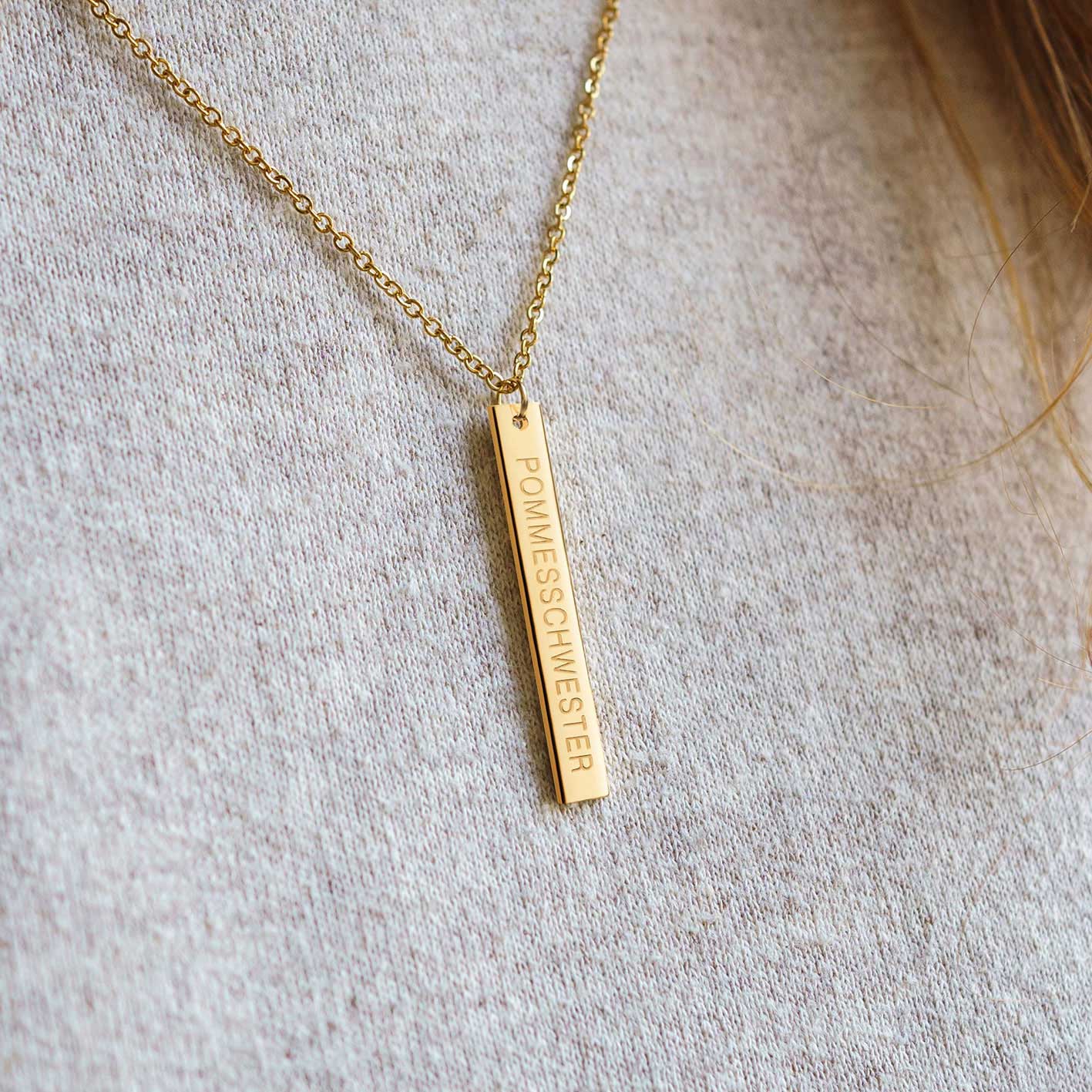 Nahaufnahme einer goldenen Kette mit länglichem Anhänger mit "Pommesschwester"-Gravur auf beigem Pulli