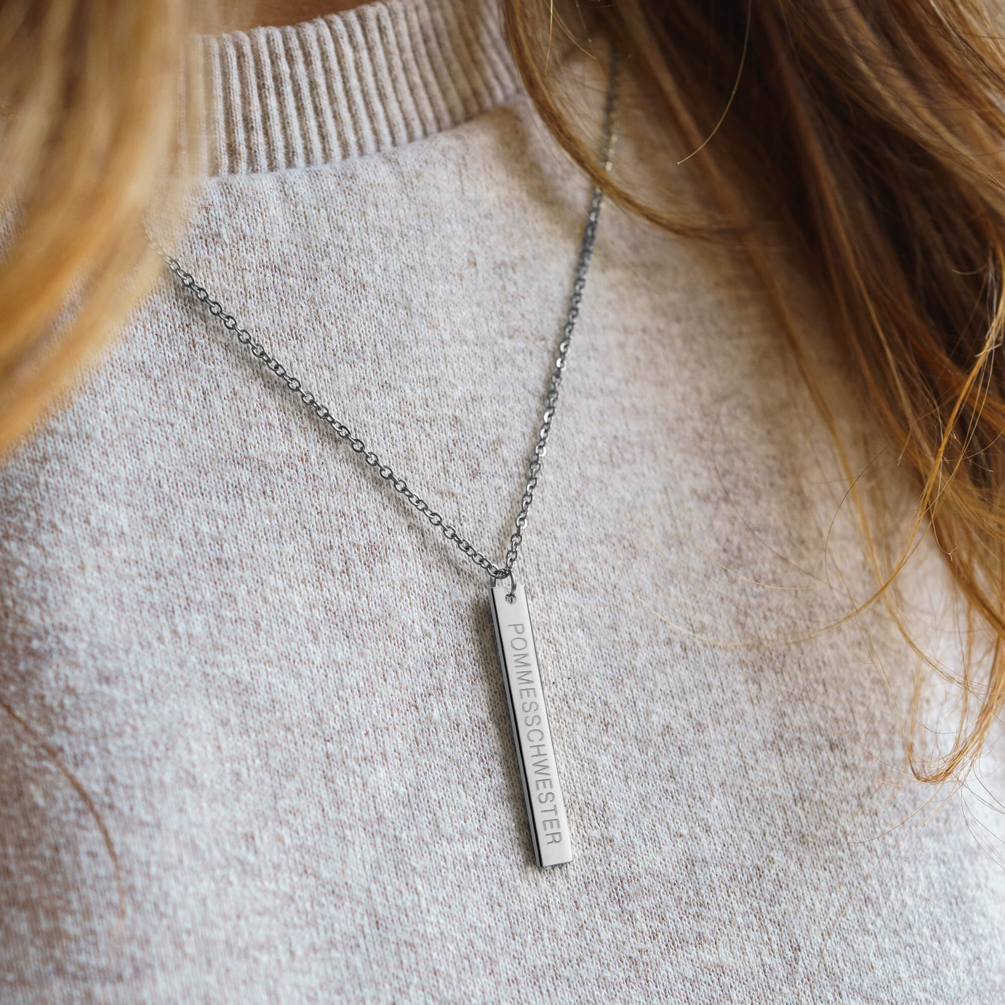 Frau mit langen Haaren trägt silberne Kette mit länglichem Anhänger mit "Pommesschwester"-Gravur über beigen Pulli.