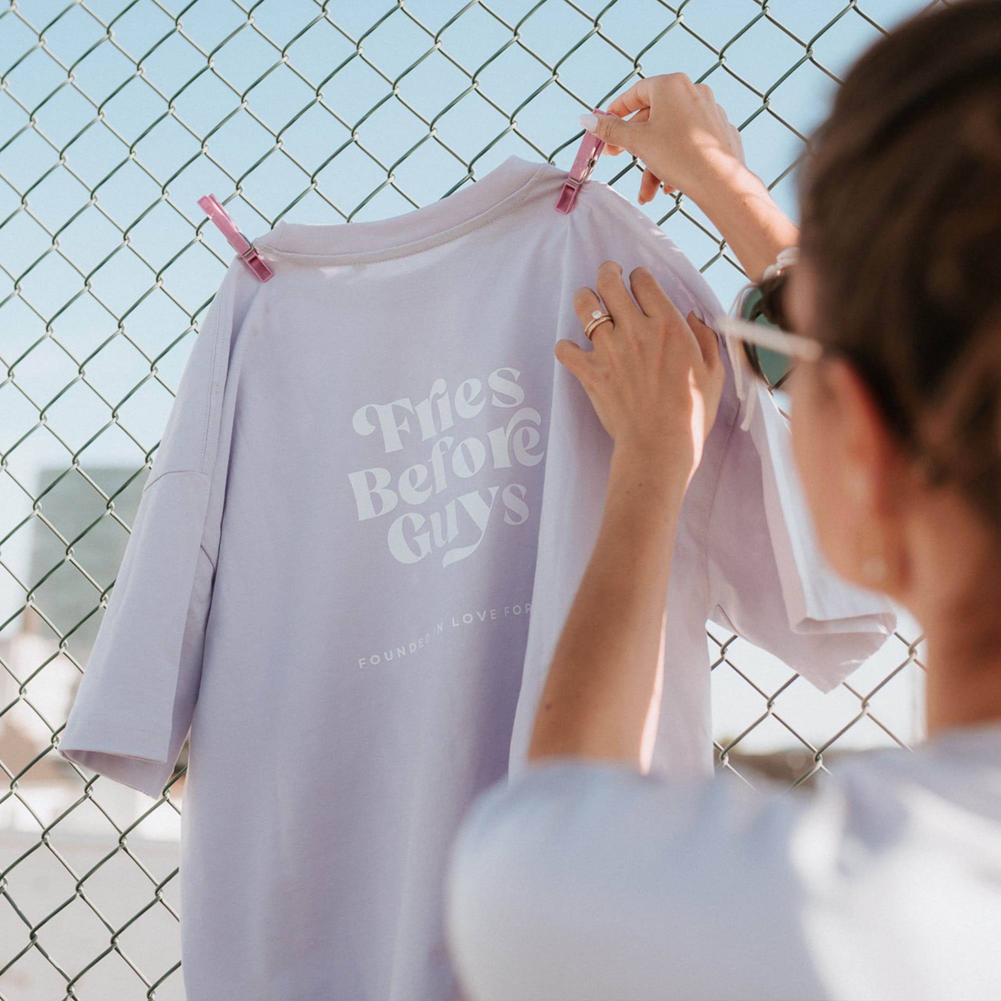 Lilanes T-Shirt mit großem weißen "Fries Before Guys"-Rückenprint wird von einer Frau mit Wäscheklammern an einem Zaun aufgehangen