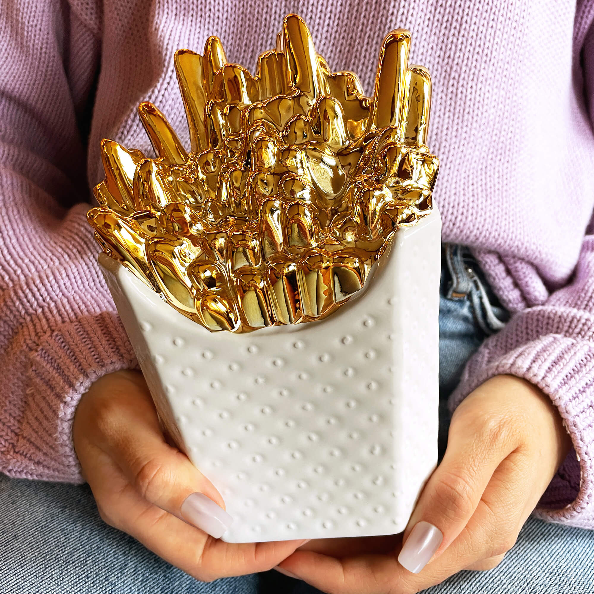 Frau hält Spardose in Form einer weißen Pommestüte mit goldenen Pommes in den Händen.