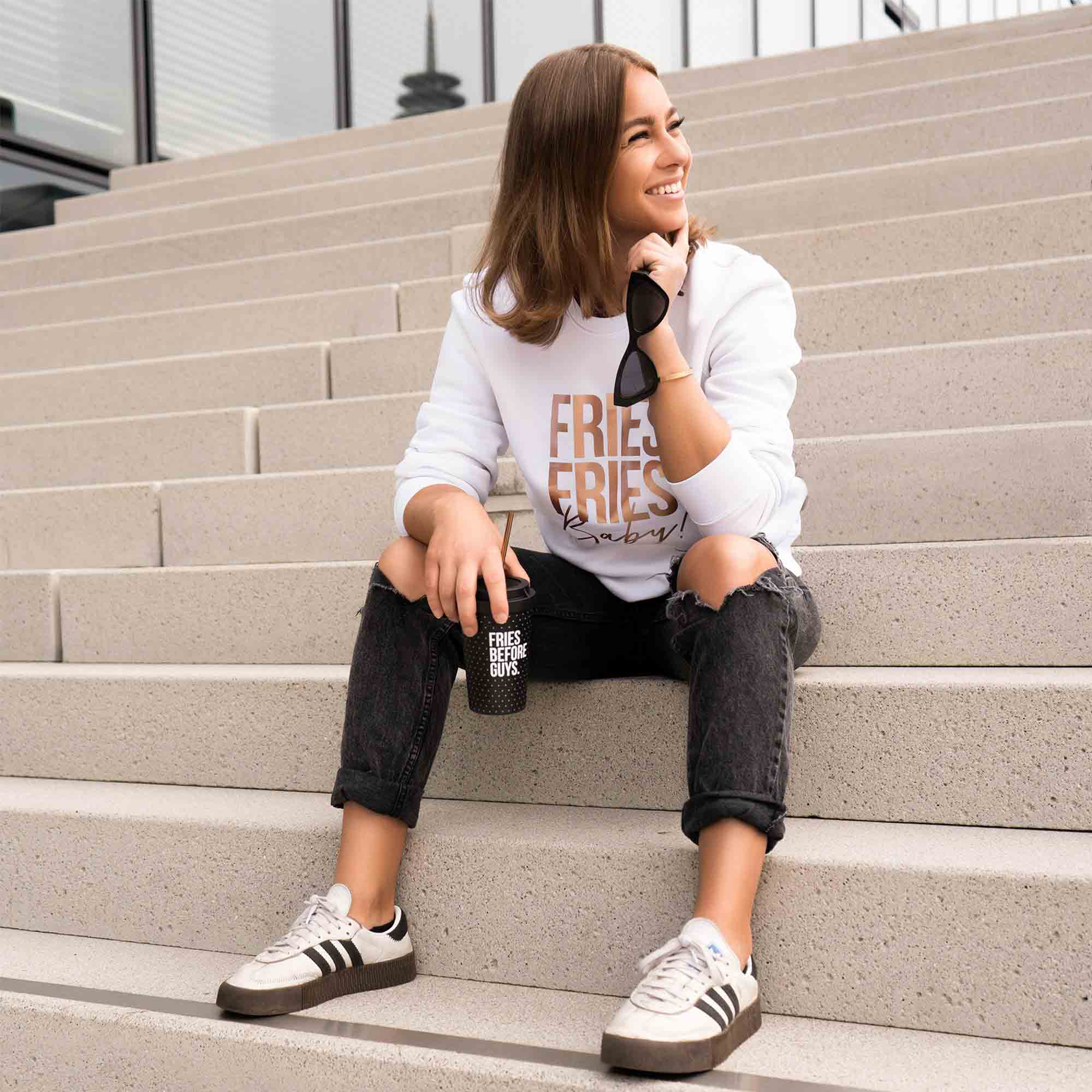 Frau trägt weißen Pullover mit metallic "Fries Fries Baby!"-Druck auf einer Treppe