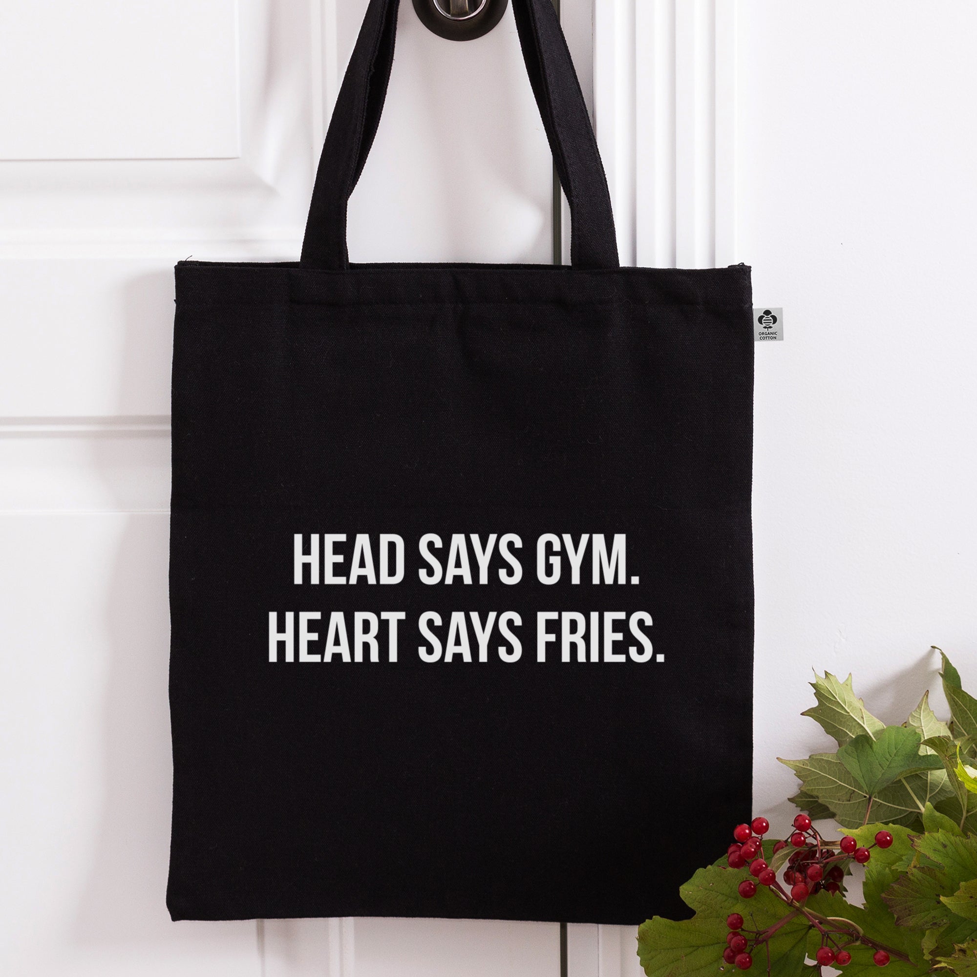 Schwarze Tragetasche mit weißem "Head Says Gym. Heart Says Fries."-Druck vor weißer Tür