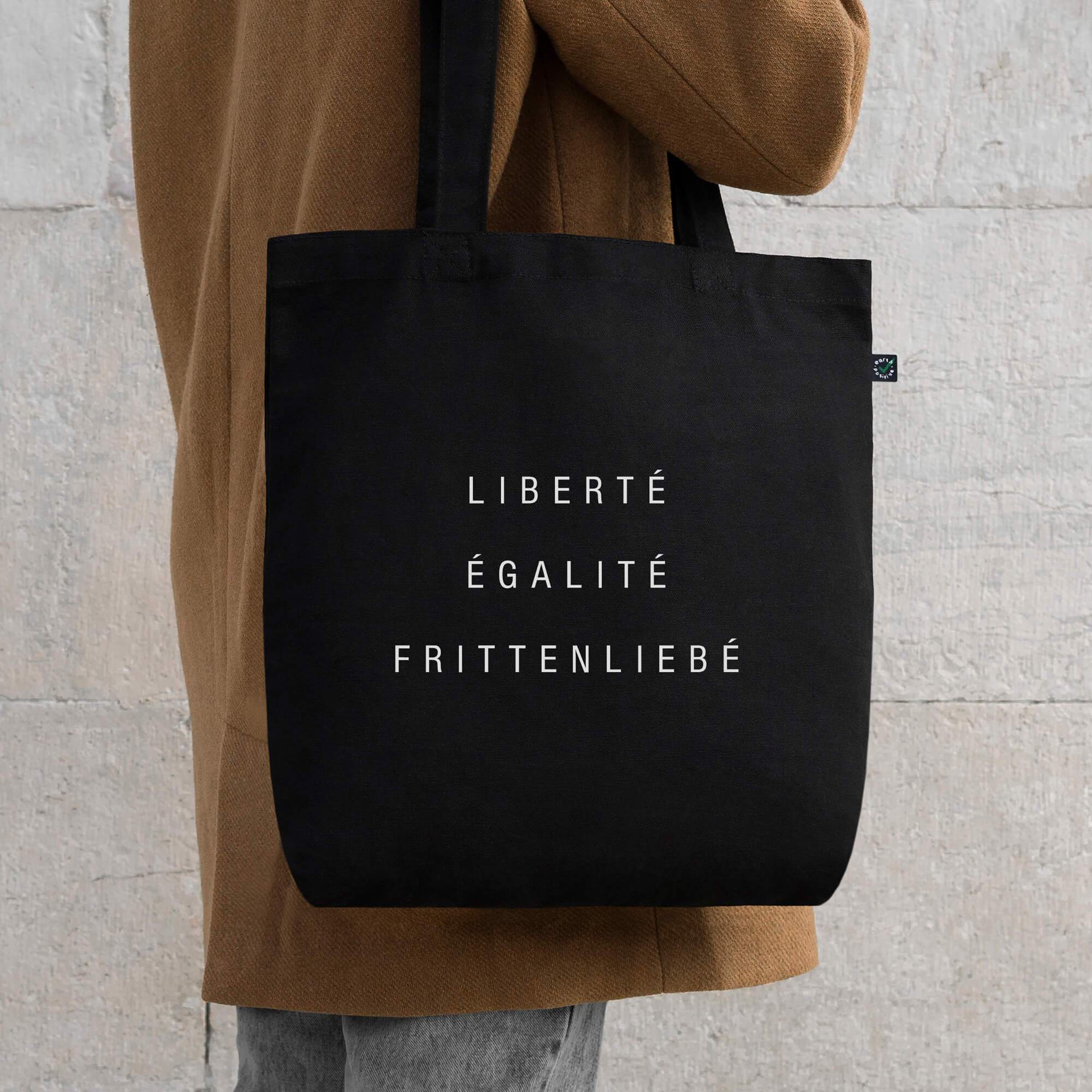 Frau in braunem Mantel trägt schwarze Tragetasche mit weißem "Liberté. Egalité. Frittenliebé"-Druck.