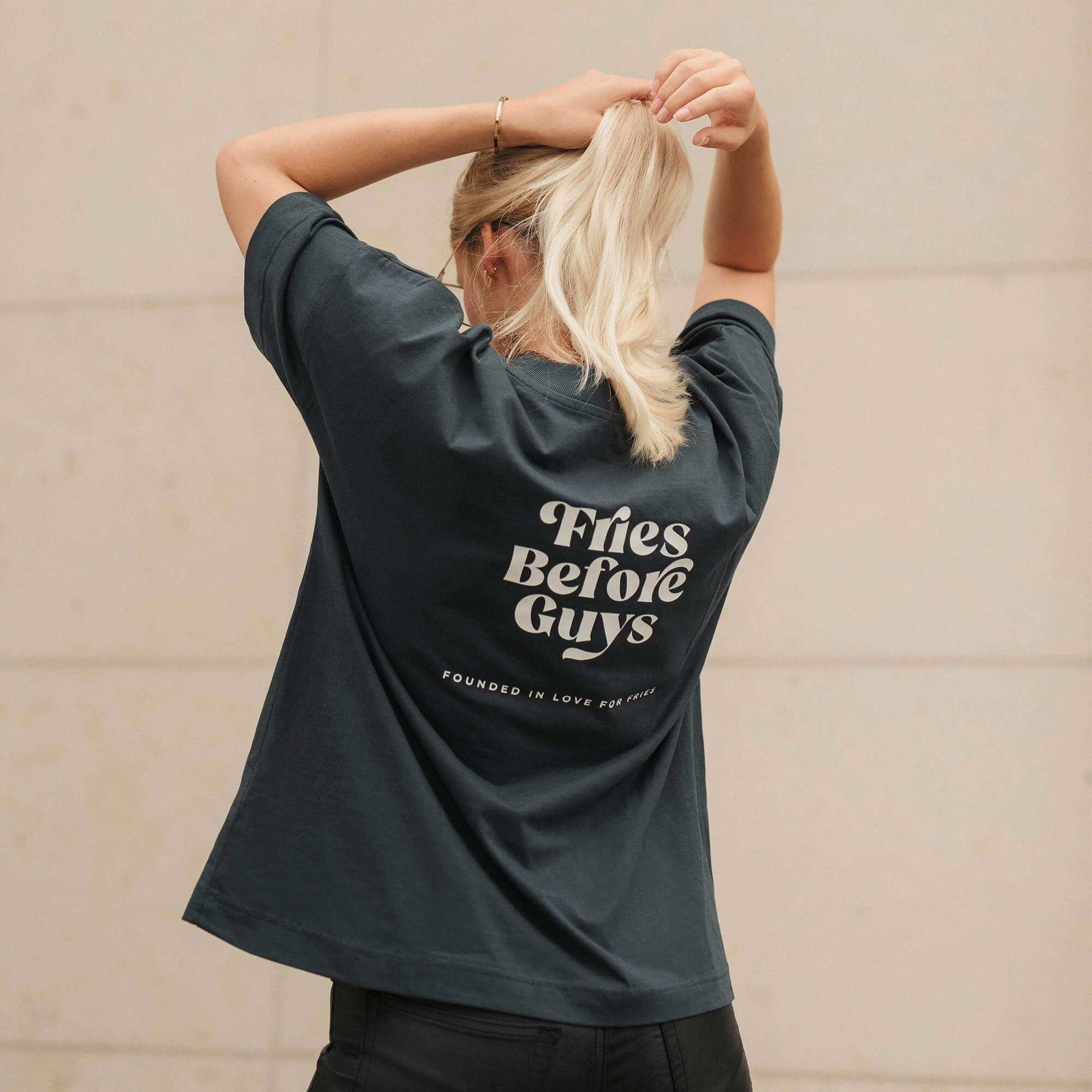 Blonde Frau trägt granitfarbenes T-Shirt mit weißem "Fries Before Guys"-Rückenprint.