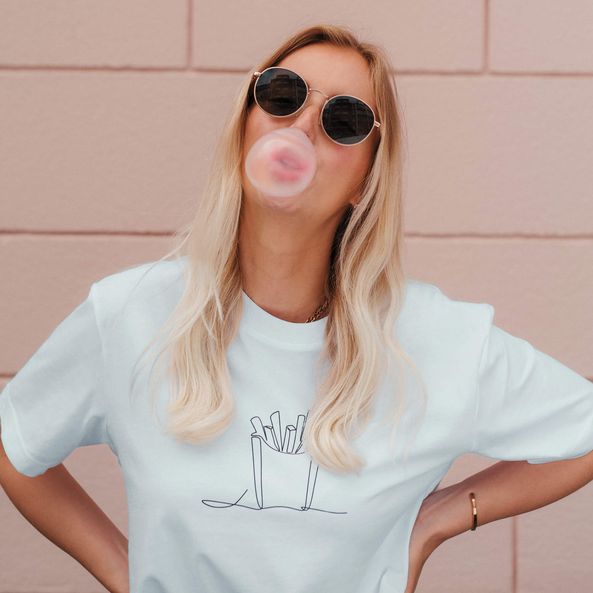 Frau vor rosaner Steinwand trägt weißes T-Shirt mit schwarzem Motiv einer Pommestüte.